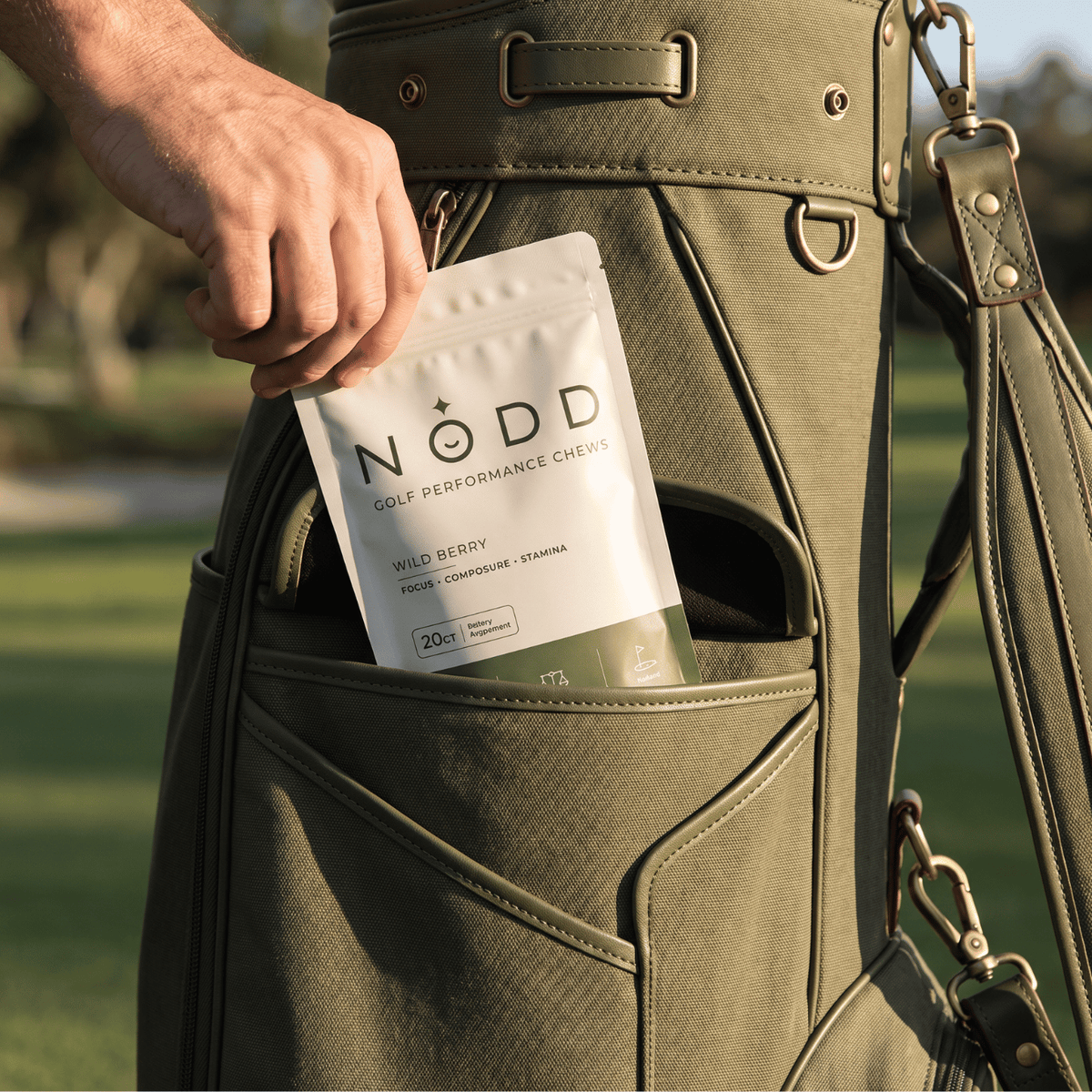 A person's hand placing a pouch of NODD Golf Performance Cheeks into the side pocket of an olive green golf bag on a golf course.